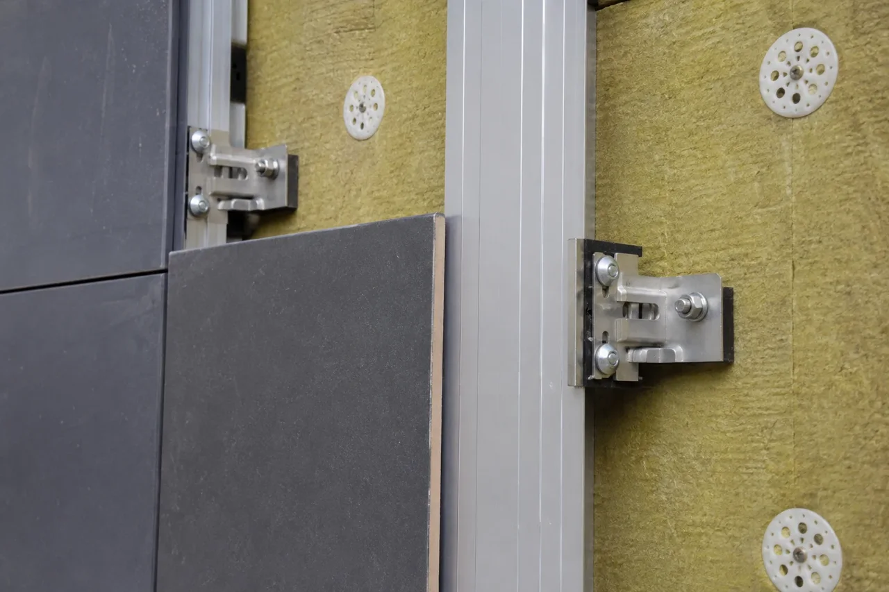 Close-up of a dark exterior cladding panel attached to vertical aluminum carrier rails with stainless steel undercut clips over mineral wool insulation
