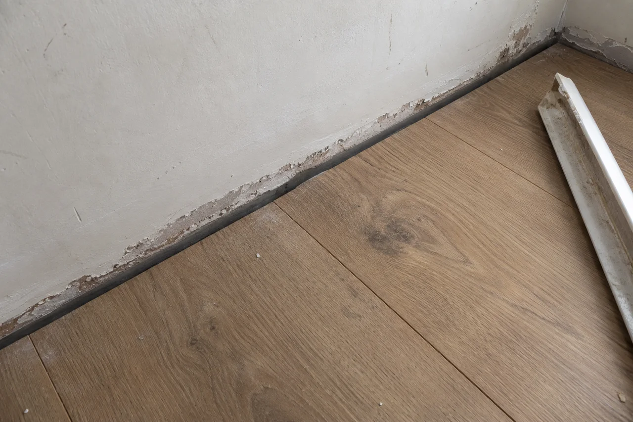 Floor planks stop beside a plaster wall, with the removed white baseboard resting to one side
