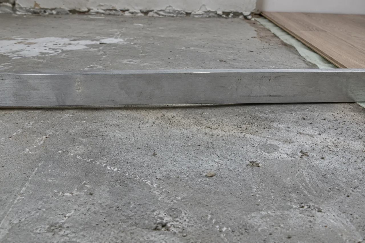 An unmarked metal straightedge rests across bare concrete beside the edge of remaining floor covering